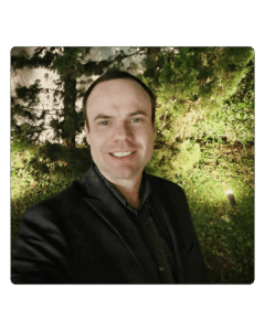 Homem sorrindo ao ar livre durante a noite, usando blazer escuro, com iluminação suave e vegetação ao fundo.